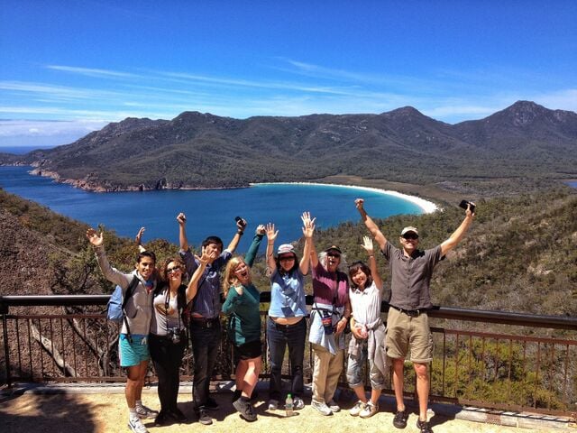 Group of travellers celebrating at Wineglass Bay lookout with turquoise waters and mountain backdrop