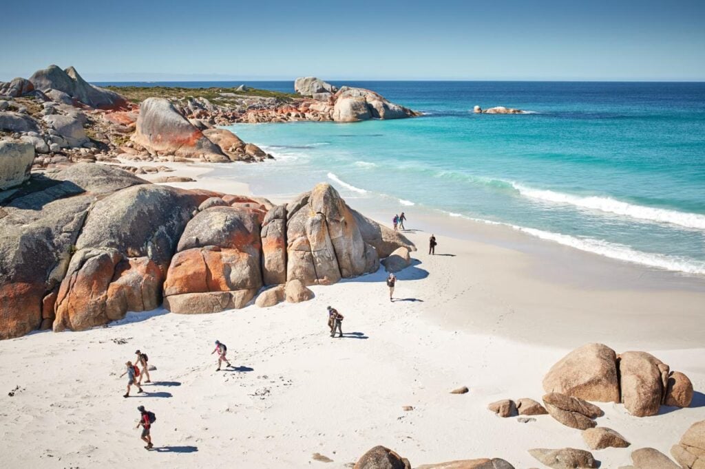 Group of walkers on white sand beach framed by orange granite rocks and turquoise waves in Bay of Fires